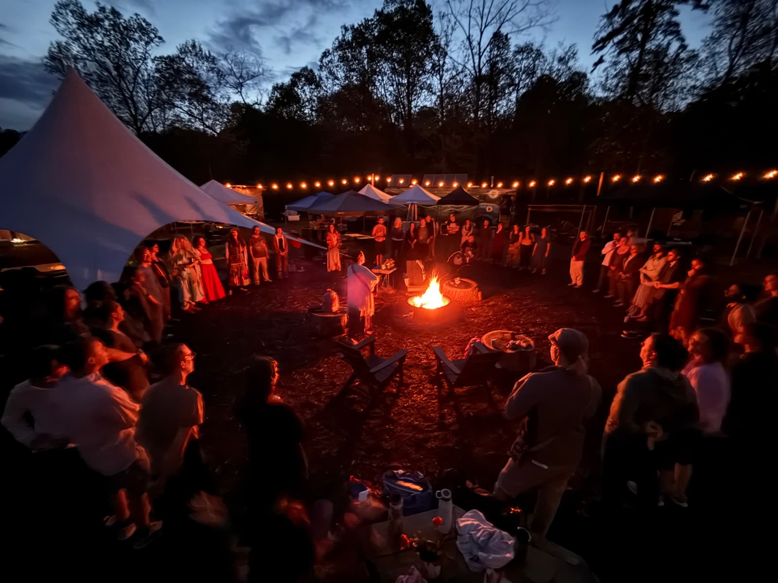 CATALY$T REVIVAL at night: a crowd standing in a circle around a fire, string lights and tents behind them